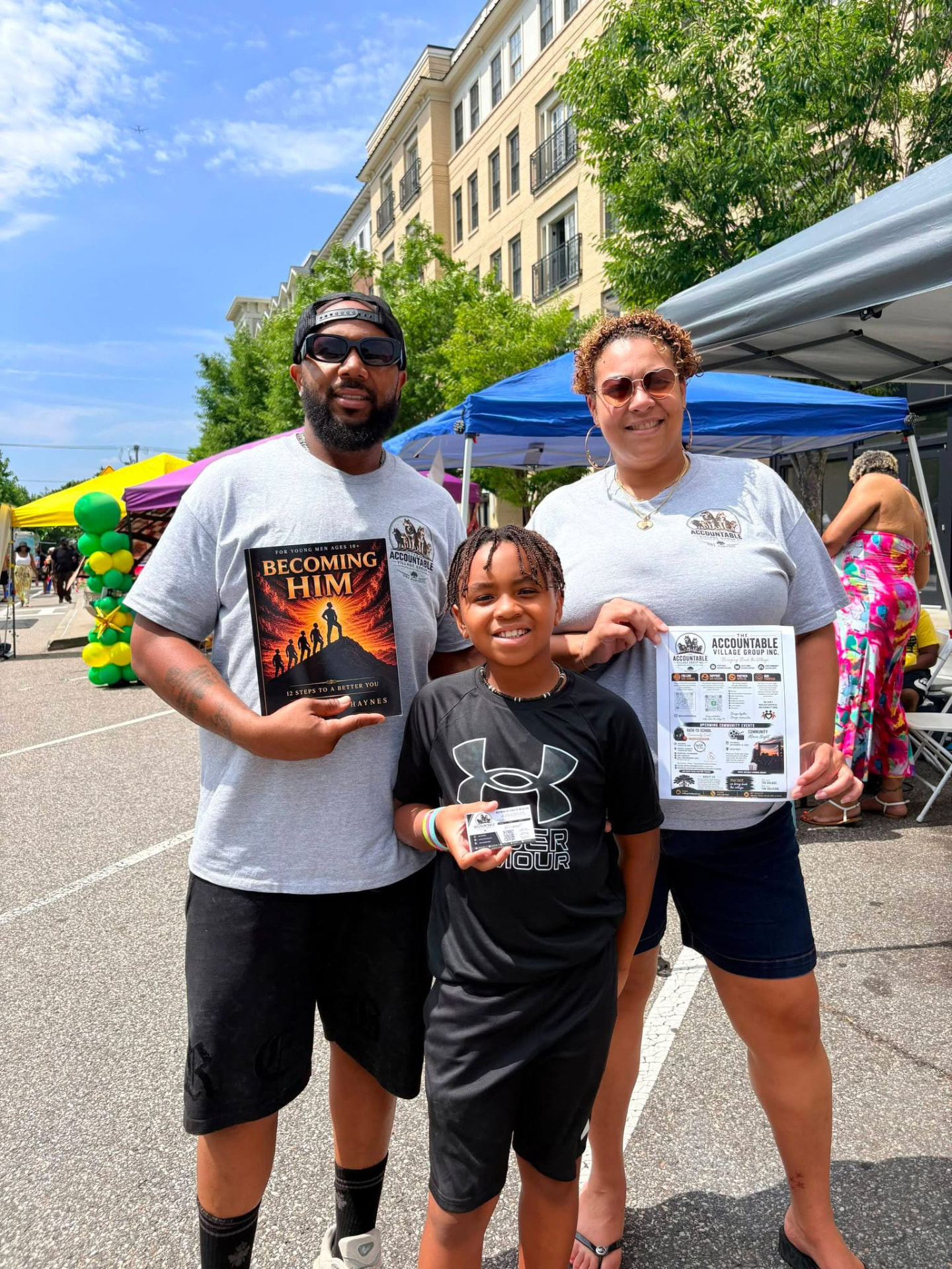 A family holding the Becoming Him book and Accountable Village flyer at a street fair